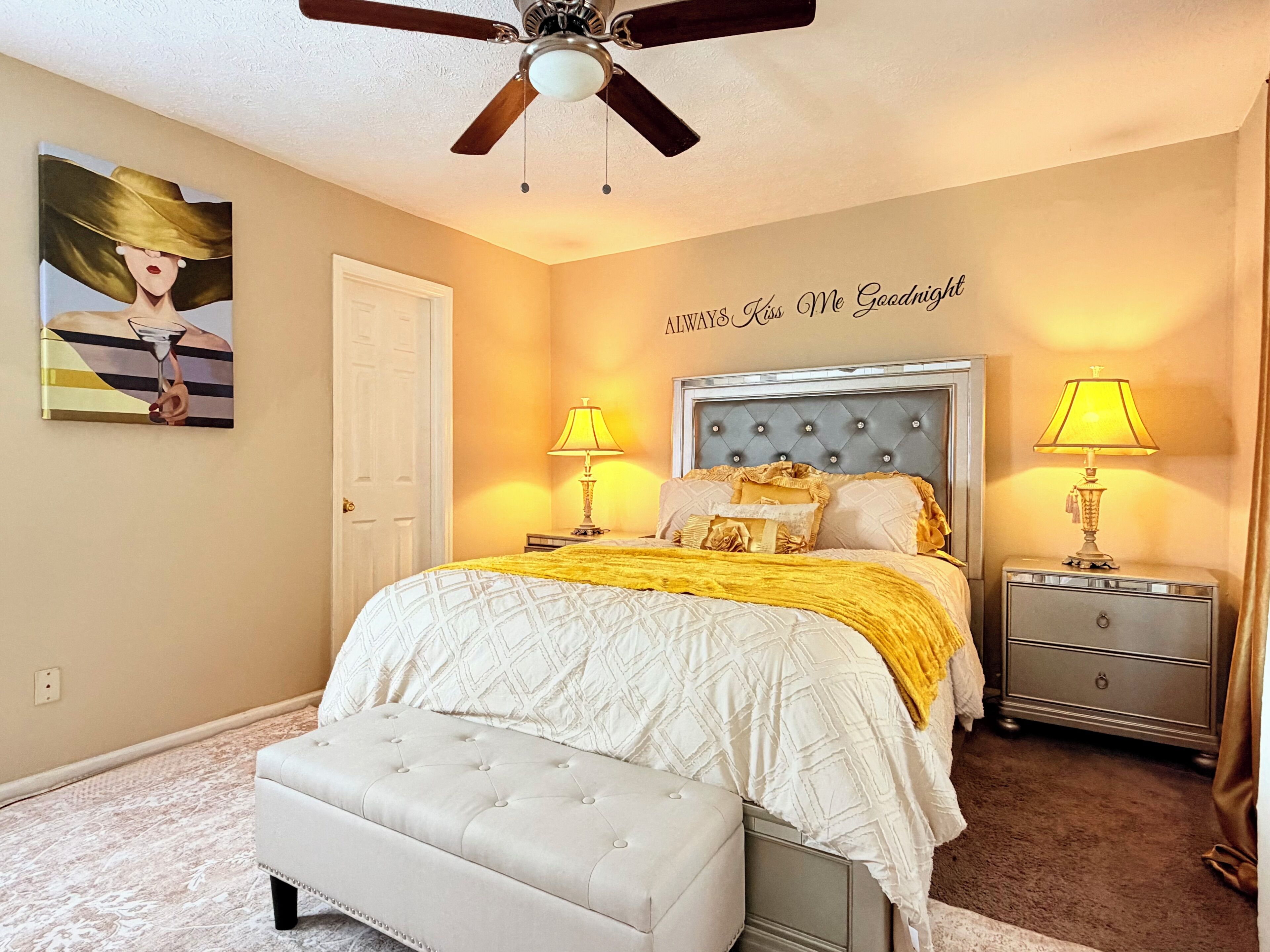A bedroom with a bed, two bedside tables with lamps, a ceiling fan, a door, and a painting on the wall.