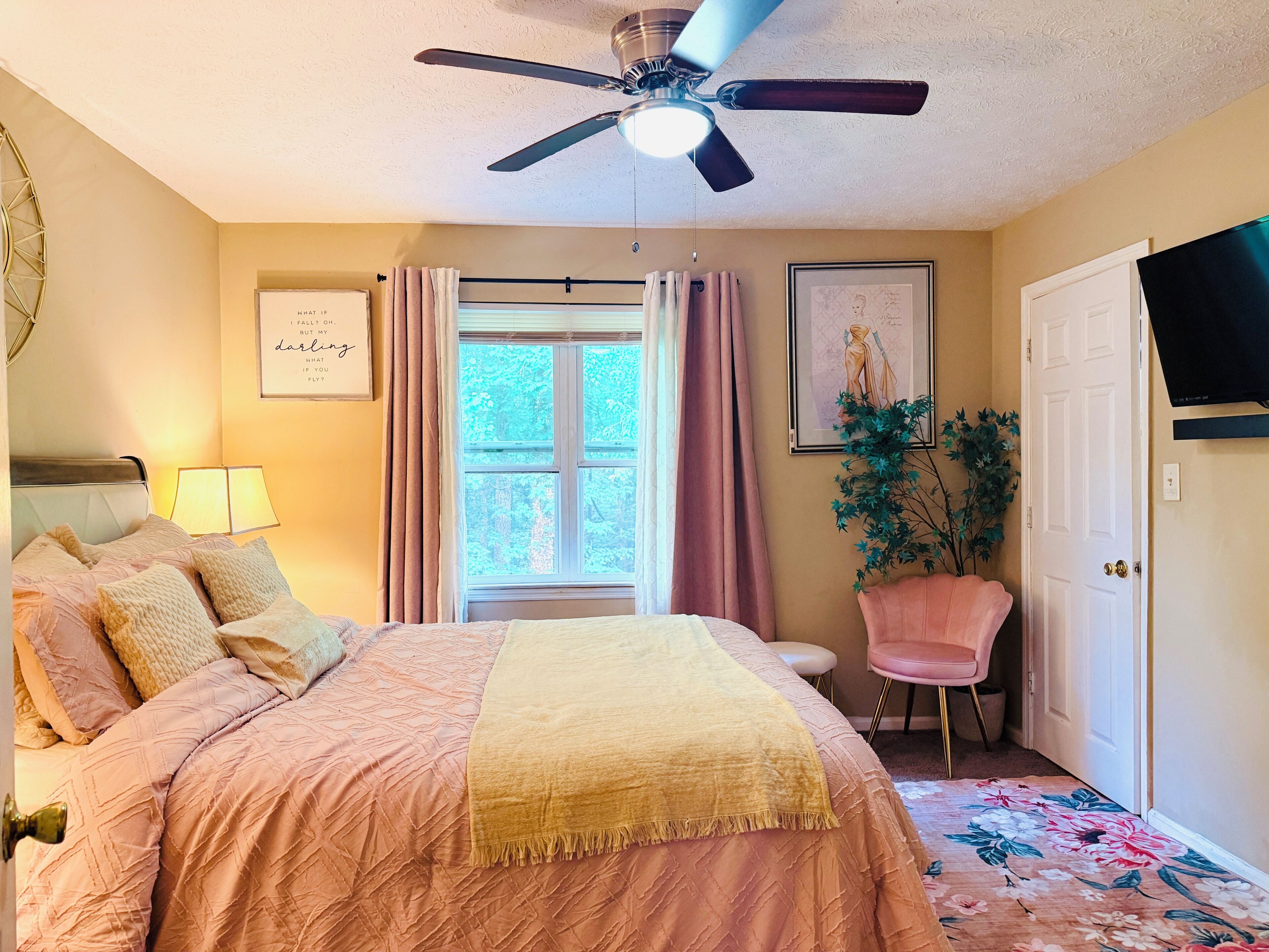 A bedroom with a bed, a ceiling fan, a window with curtains, a chair, a plant, and a TV.