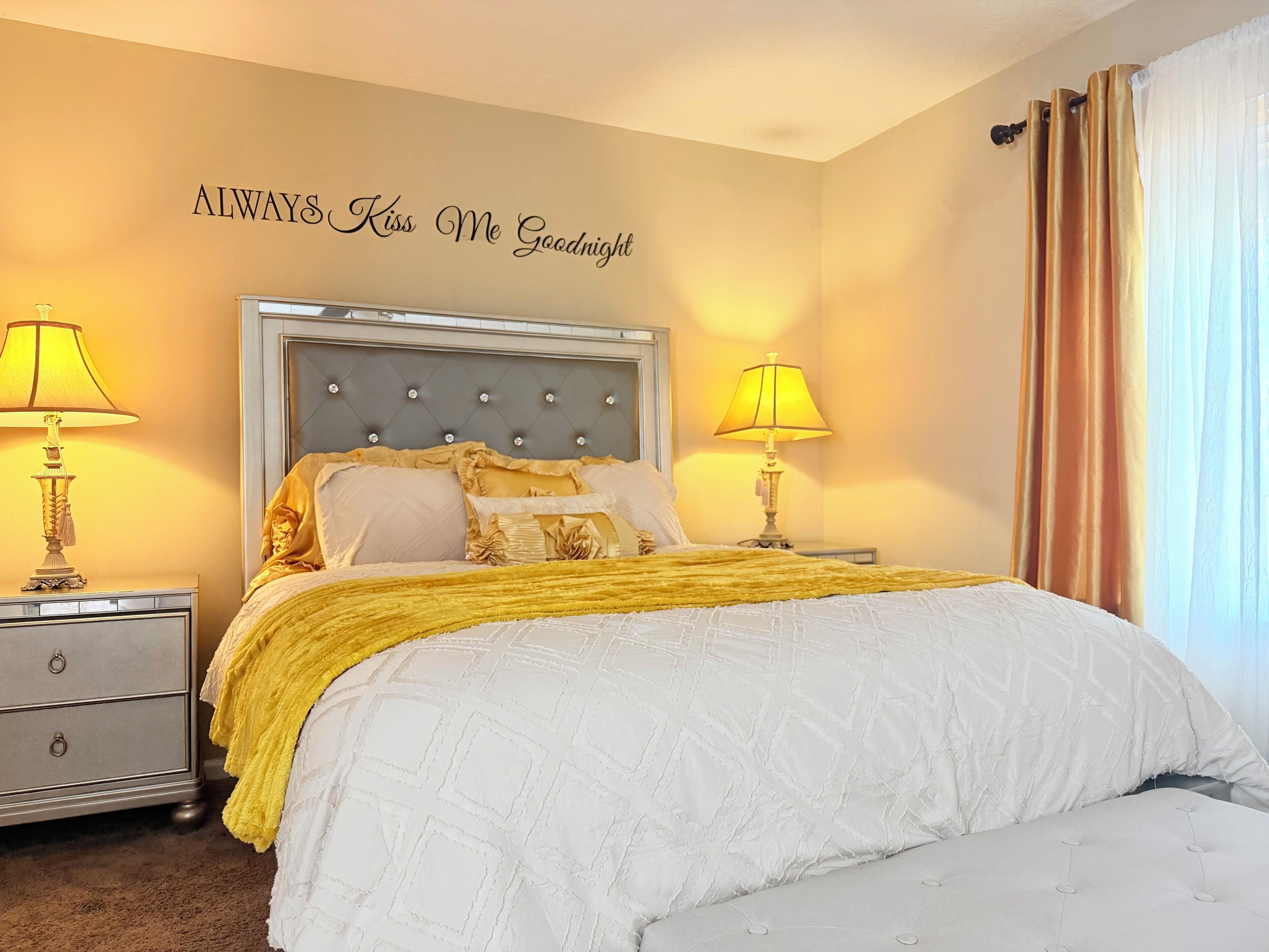 A bedroom with a bed, two bedside tables with lamps, a decorative wall sign, and a window with curtains.