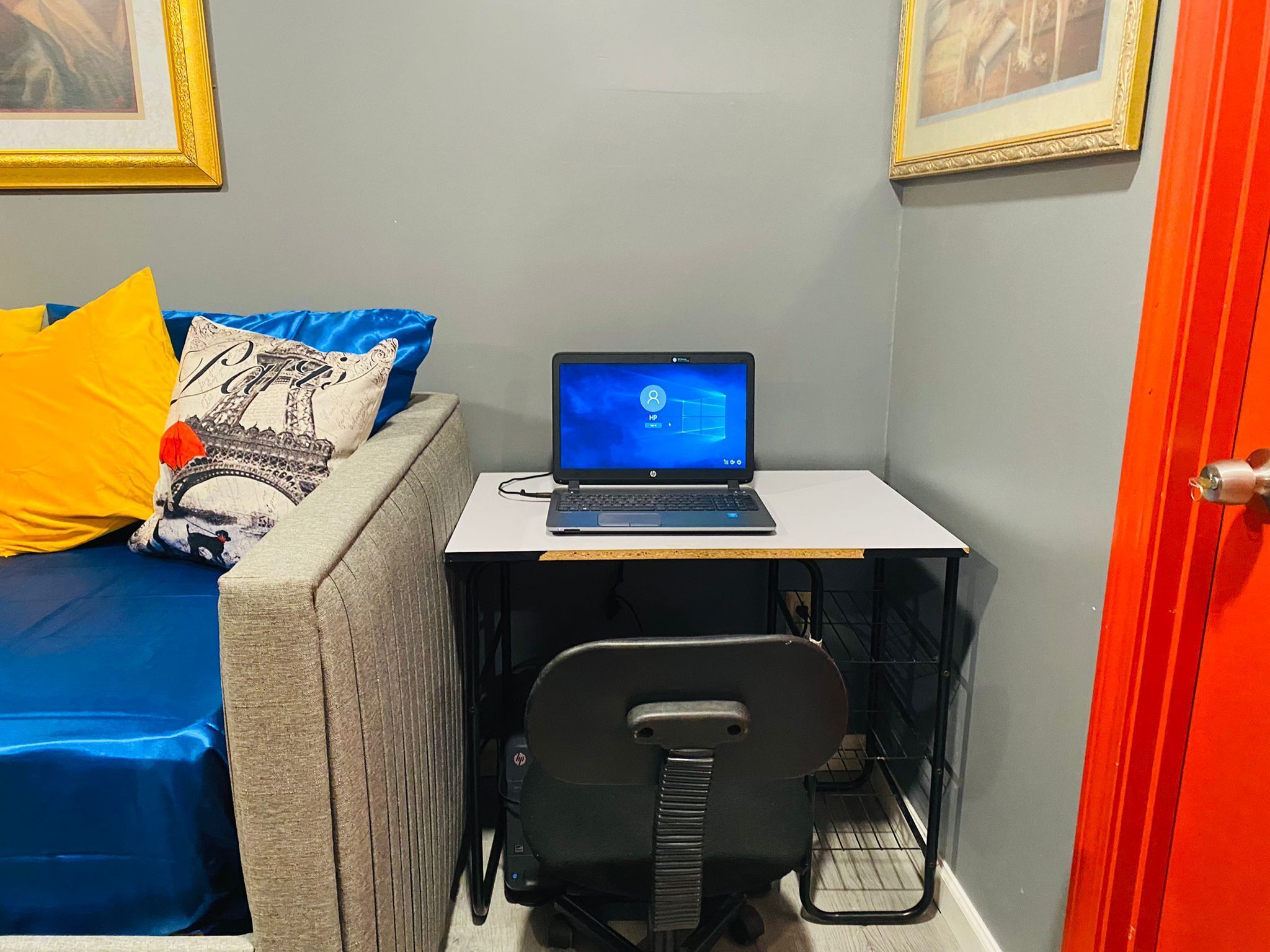 A small room with a laptop on a desk, a chair, a bed with pillows, and a door.