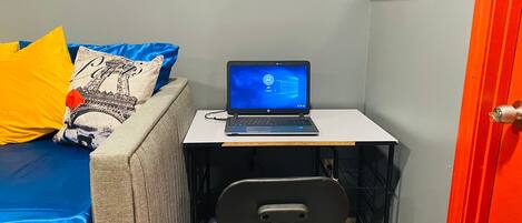 A small room with a laptop on a desk, a chair, a bed with pillows, and a door.