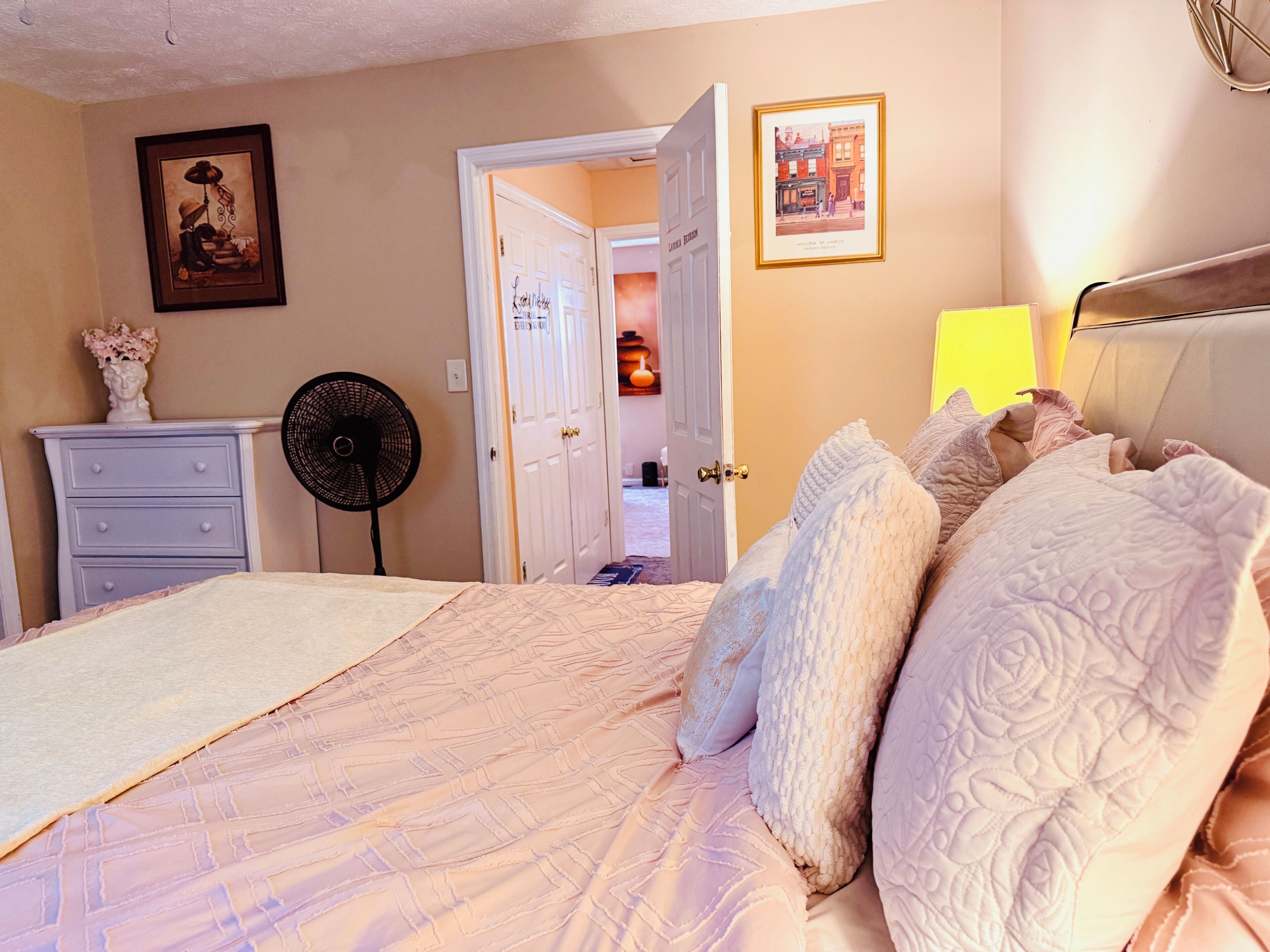 A bedroom with a bed, a dresser, a fan, and a door leading to another room.