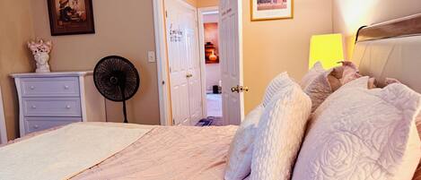 A bedroom with a bed, a dresser, a fan, and a door leading to another room.