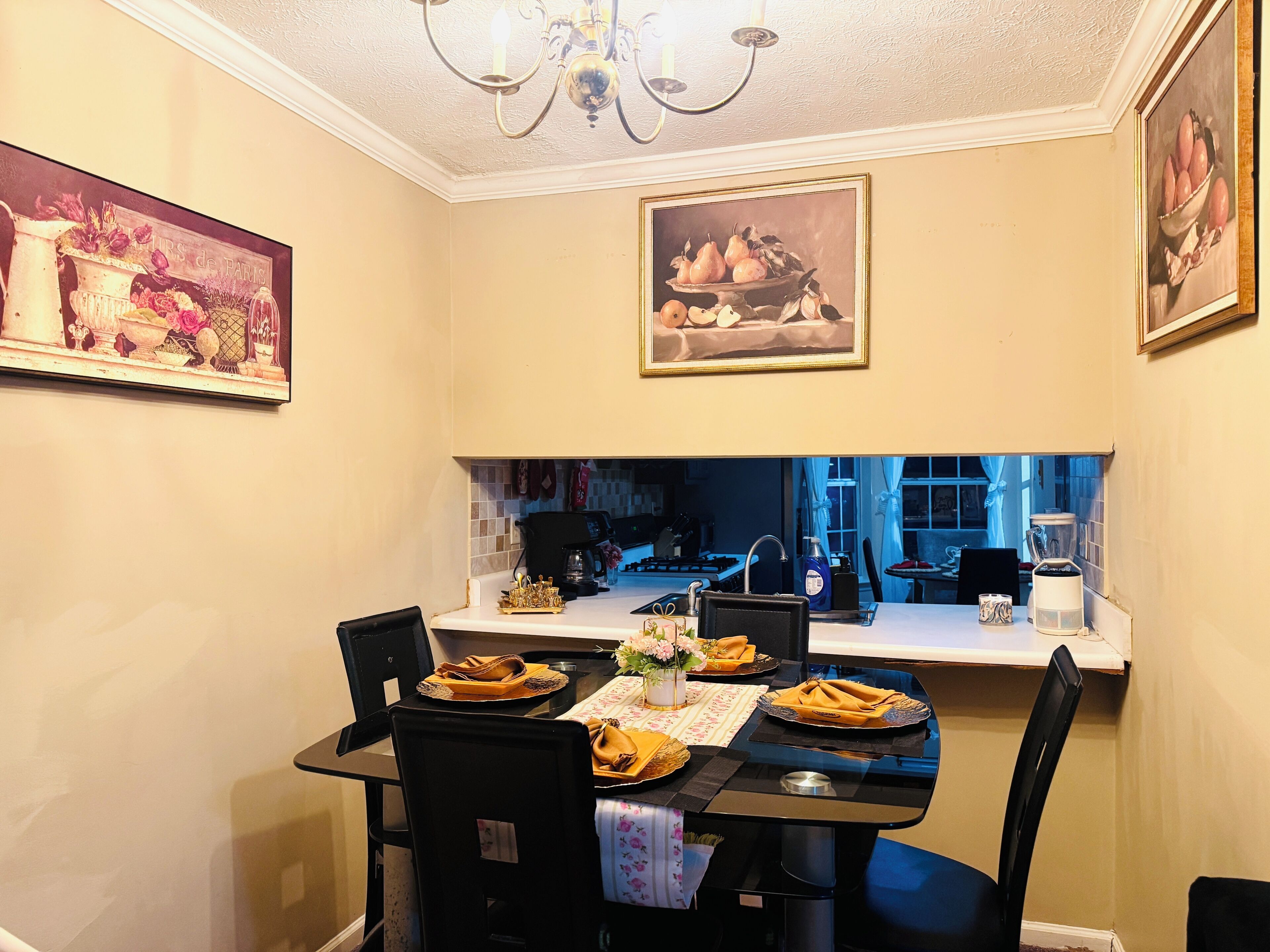 Un comedor con una mesa dispuesta para cuatro personas, una araña y cuadros en las paredes.
