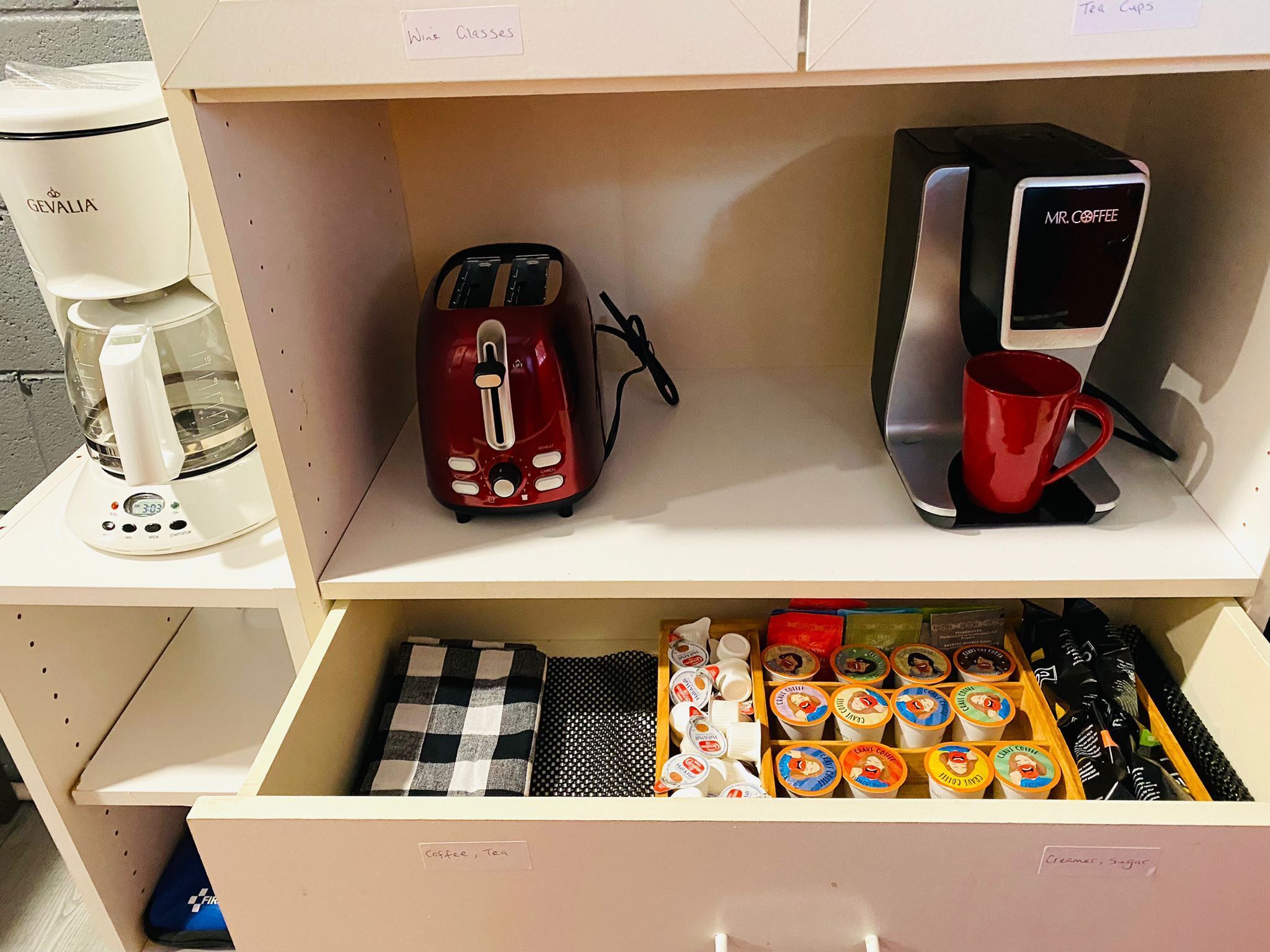 Une petite cuisine équipée d’une machine à café, d’un grille-pain et d’un appareil à thé, le tout posé sur le plan de travail. En dessous, se trouve un tiroir contenant différents types de capsules de café et de thé.