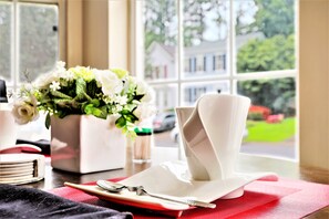 A neatly arranged table with a white napkin holder, a spoon, and a red placemat. A potted plant with white flowers is visible in the background. - Sweet Home Georgia (Norcross)