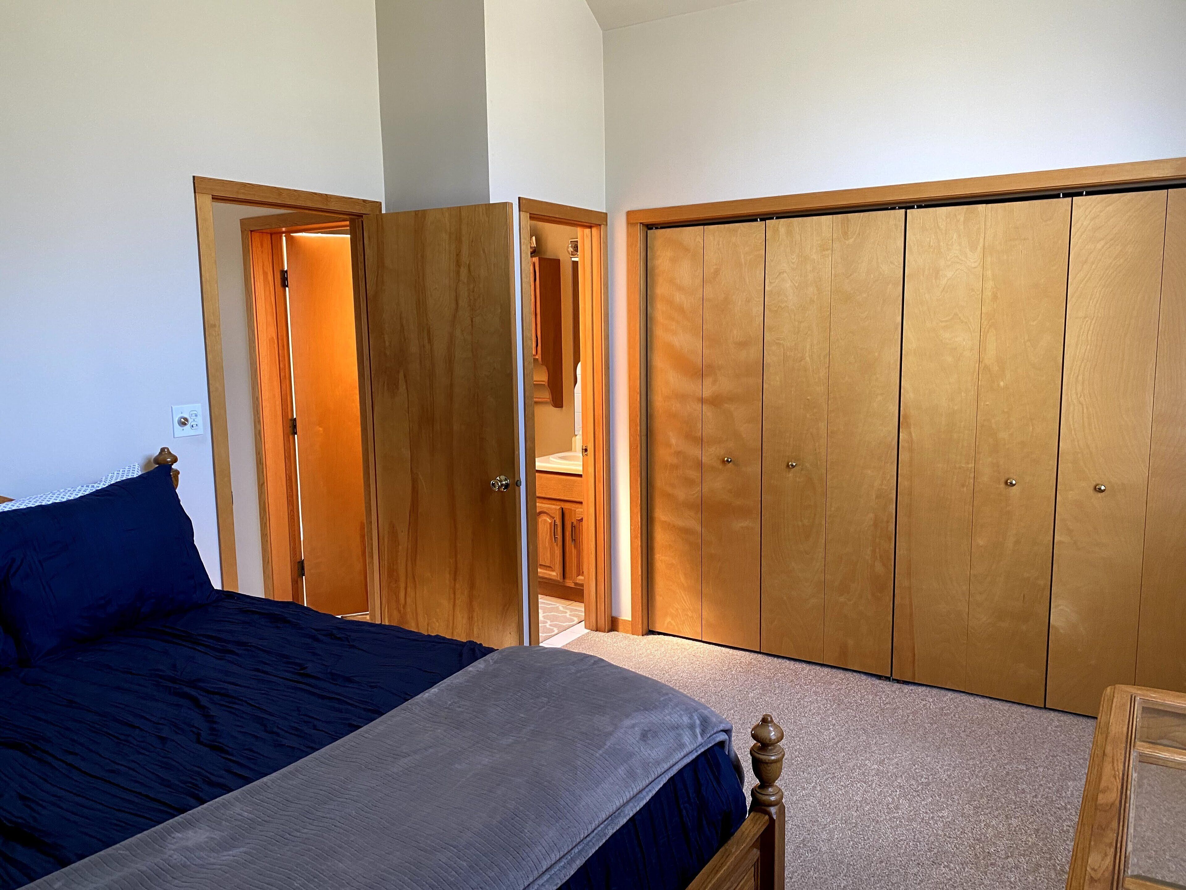 Un dormitorio con cama, armario de madera y baño visible a través de una puerta abierta.