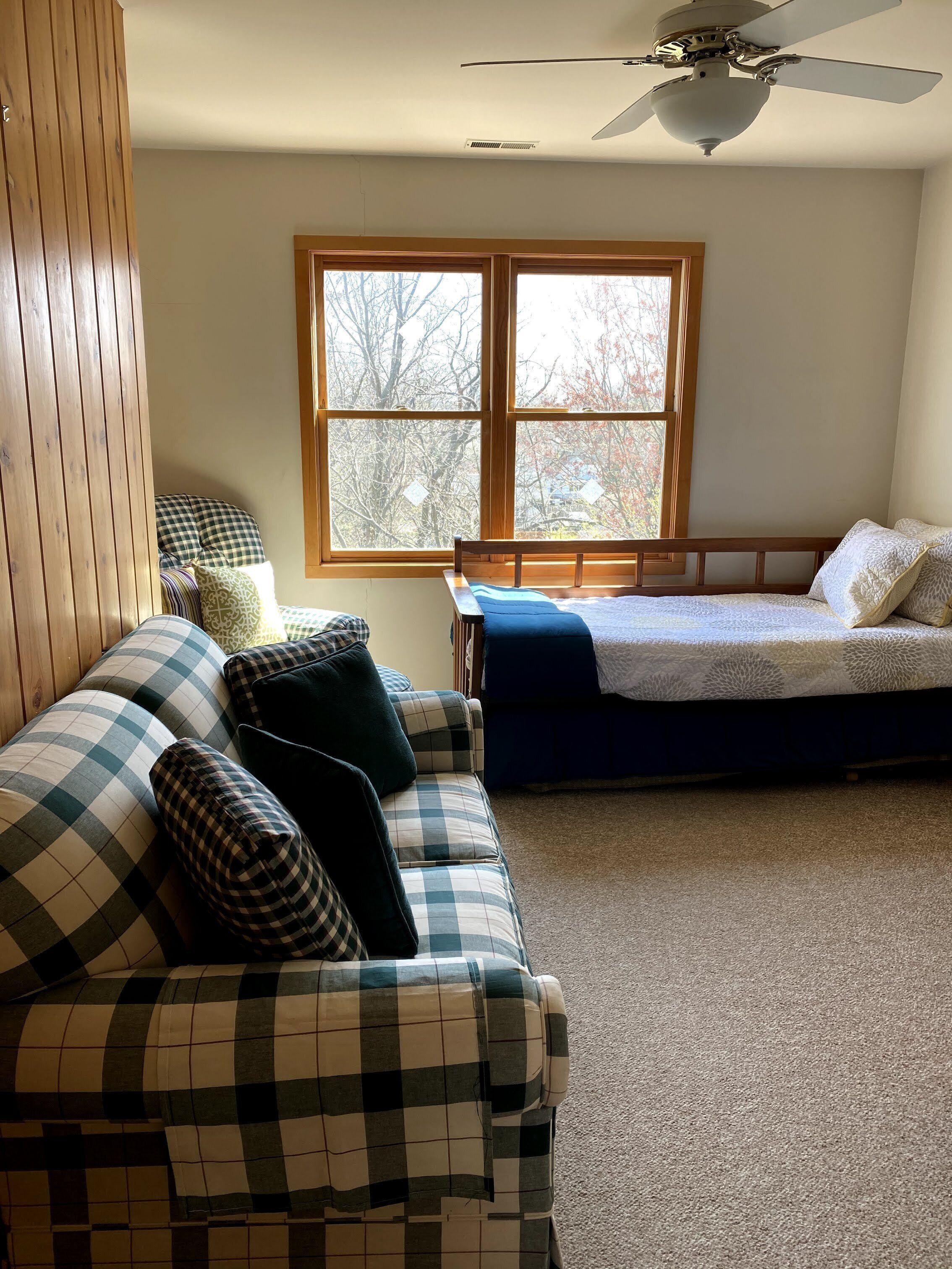 Una habitación con sofá a cuadros, cama, ventilador de techo y ventana con vista a árboles.