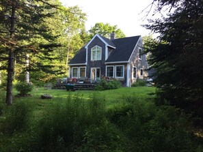 A two-story house with a steeply pitched roof, surrounded by dense greenery and trees. - Cozy Quiet Cottage in the woods, close to ocean and local activities (St George)