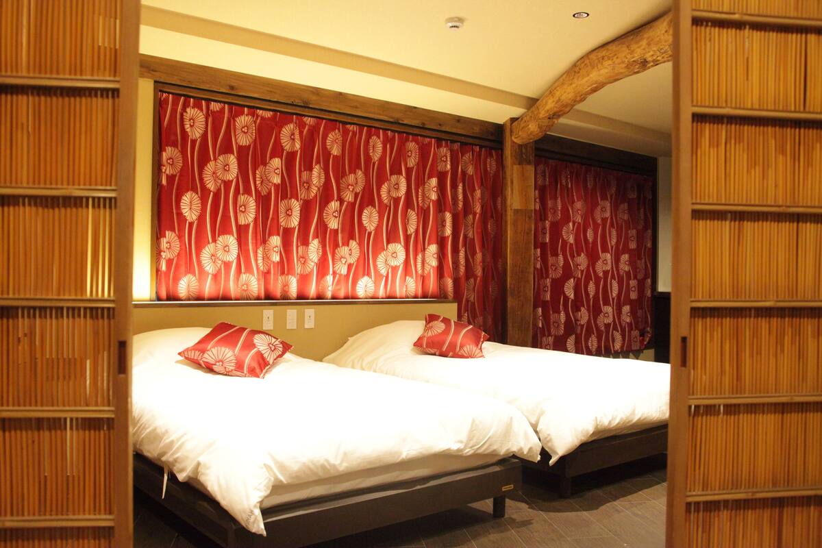 A hotel room with two beds, red floral curtains, wooden paneling, and a wooden ceiling beam.