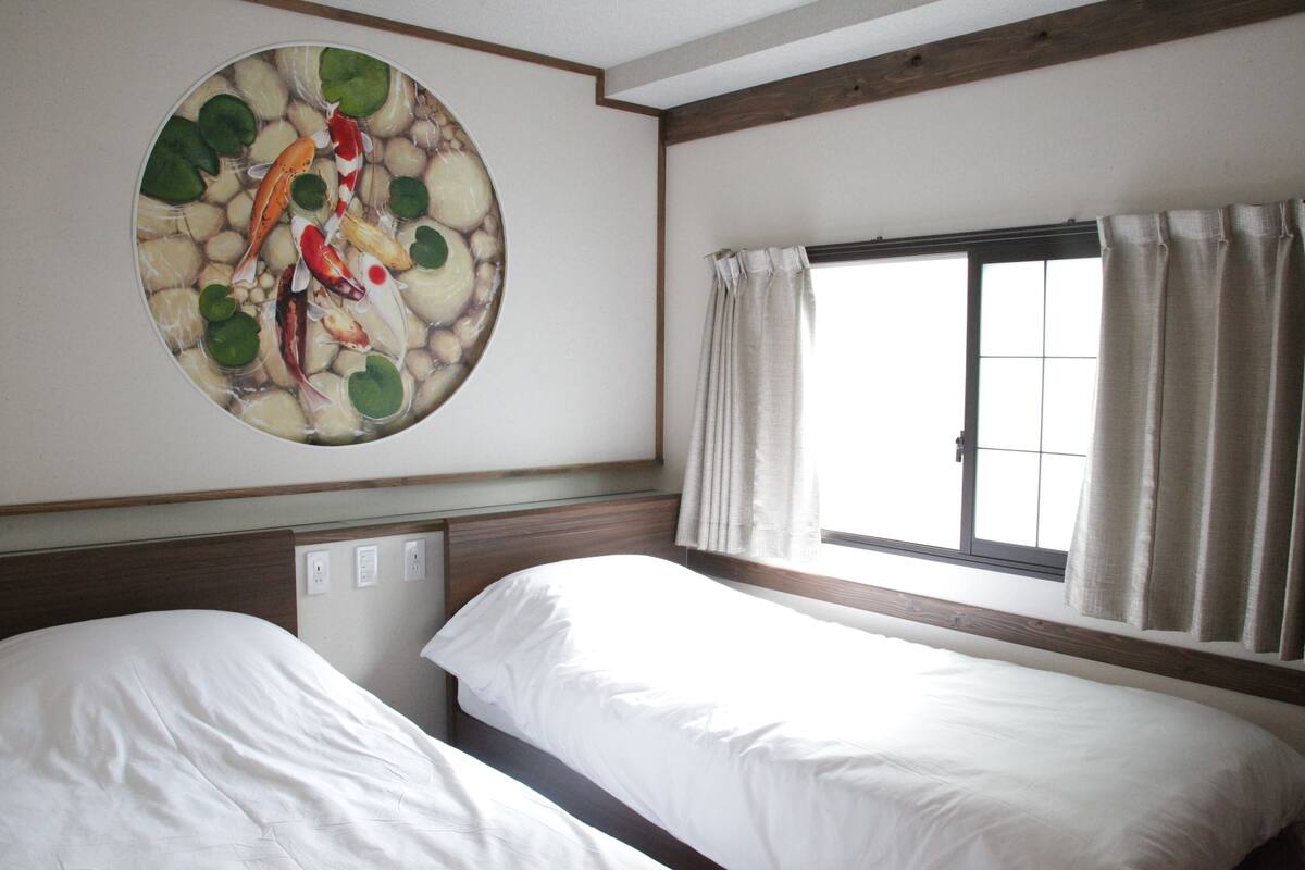 A hotel room with two beds, a large circular artwork on the wall, a window with curtains, and a wooden ceiling beam.
