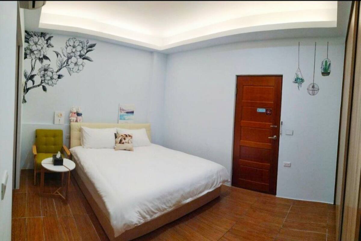 A bedroom with a bed, a green armchair, a small table, a wooden door, and a wall with a floral mural.