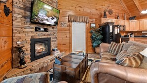 Interior acollidor d'una cabana amb una llar de pedra, un televisor penjat a la paret, armaris de fusta i una zona de seients confortable.