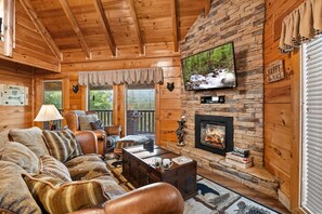 A cozy cabin interior with a fireplace, wooden walls, a television mounted above it, and a comfortable seating area with a sofa and armchairs.