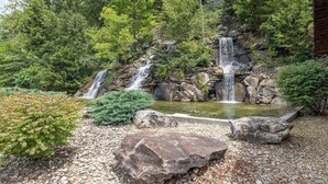 Una cascada que flueix sobre roques a un estany, envoltada de vegetació densa i una estructura de fusta.