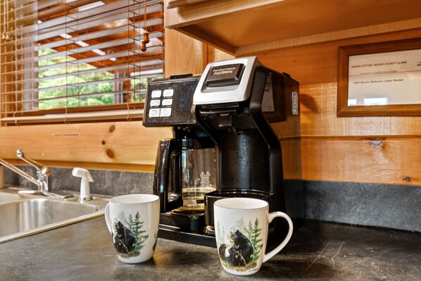 A coffee maker with a control panel, two mugs with bear designs, a sink, and a window with blinds.