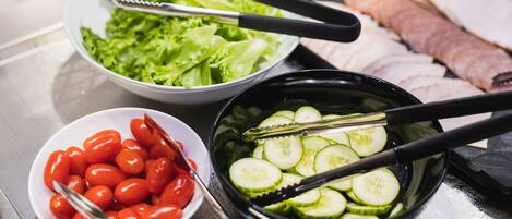 A kitchen counter with various food items including a bowl of lettuce, a bowl of cherry tomatoes, a bowl of sliced cucumbers, and a tray of sliced meat.