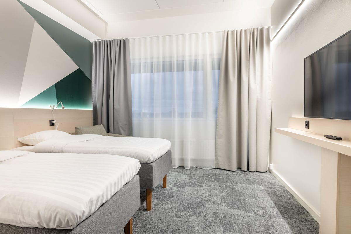 A modern hotel room with two beds, a large window with sheer curtains, a flat-screen TV, and a carpeted floor.