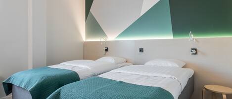 A hotel room with two beds, each with a teal blanket, a small wooden stool, and a geometric-patterned wall design.