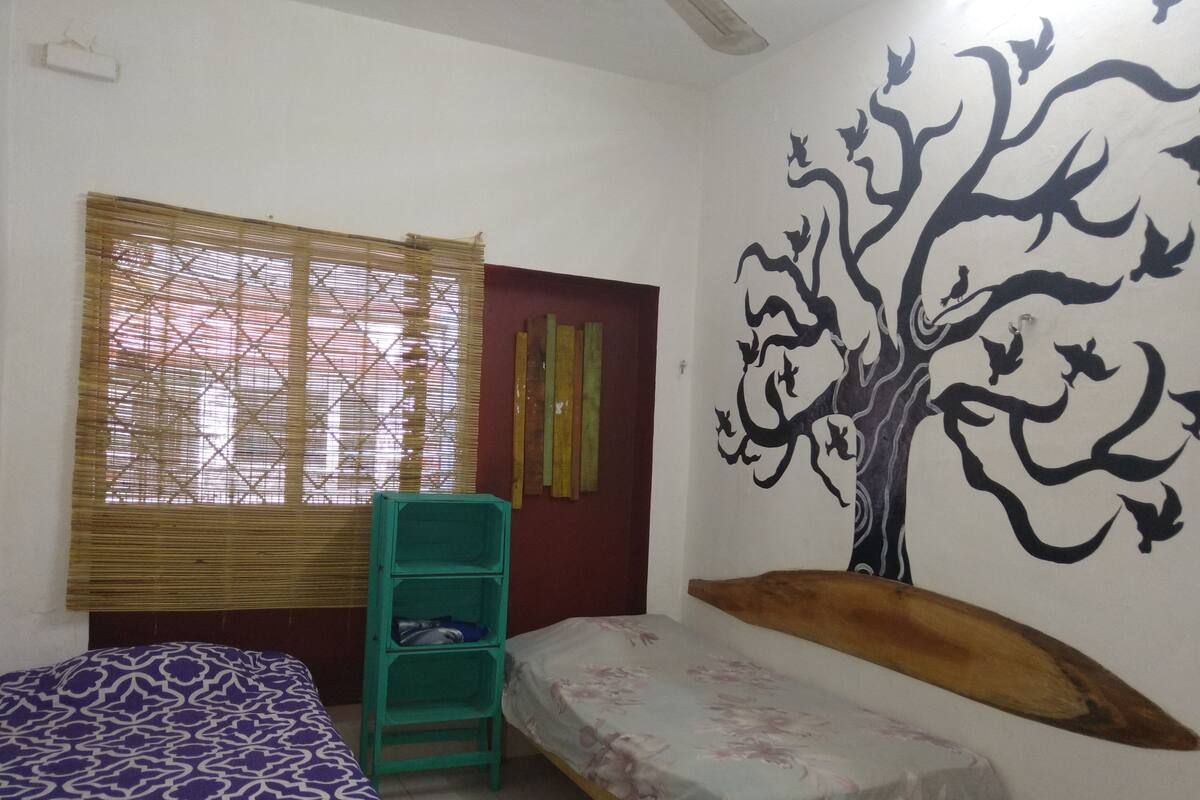 Une chambre avec deux lits, une fenêtre avec des stores en bambou, une fresque murale et une étagère verte.