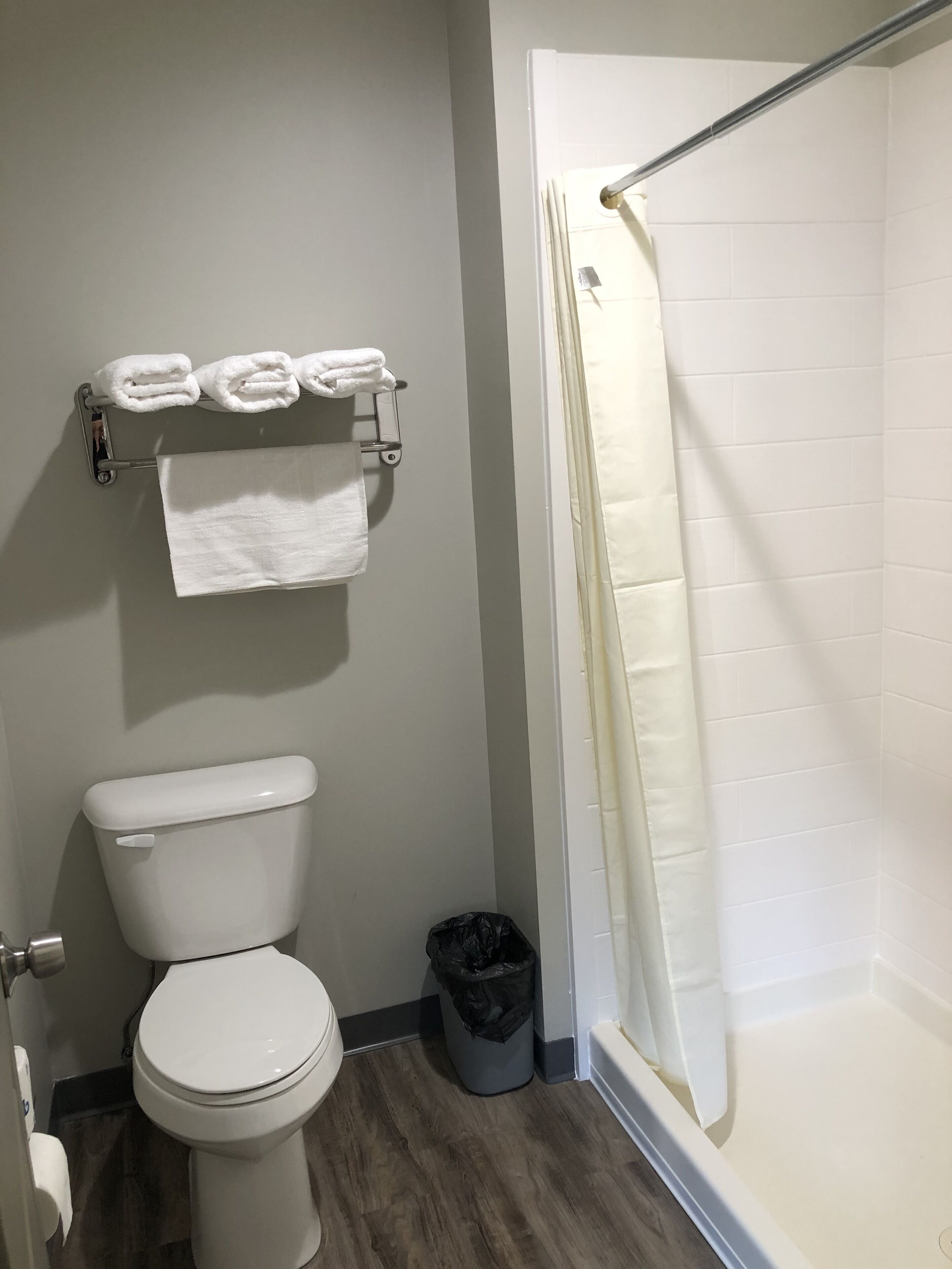 A bathroom with a toilet, a towel rack with folded towels, a trash can, and a shower area with a curtain.