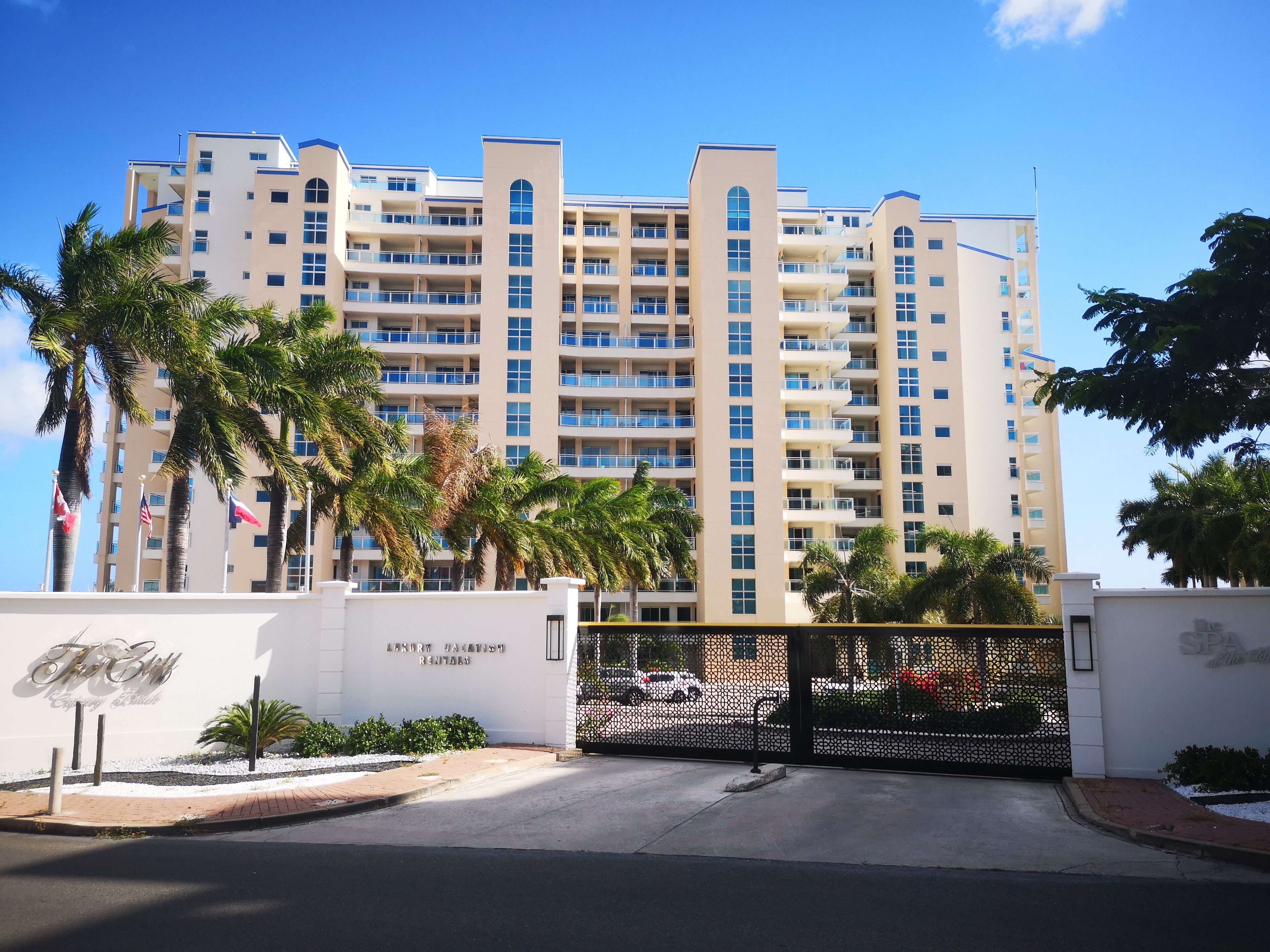 A multi-story hotel with a modern design, featuring balconies on each floor, a gated entrance, and palm trees in the foreground.