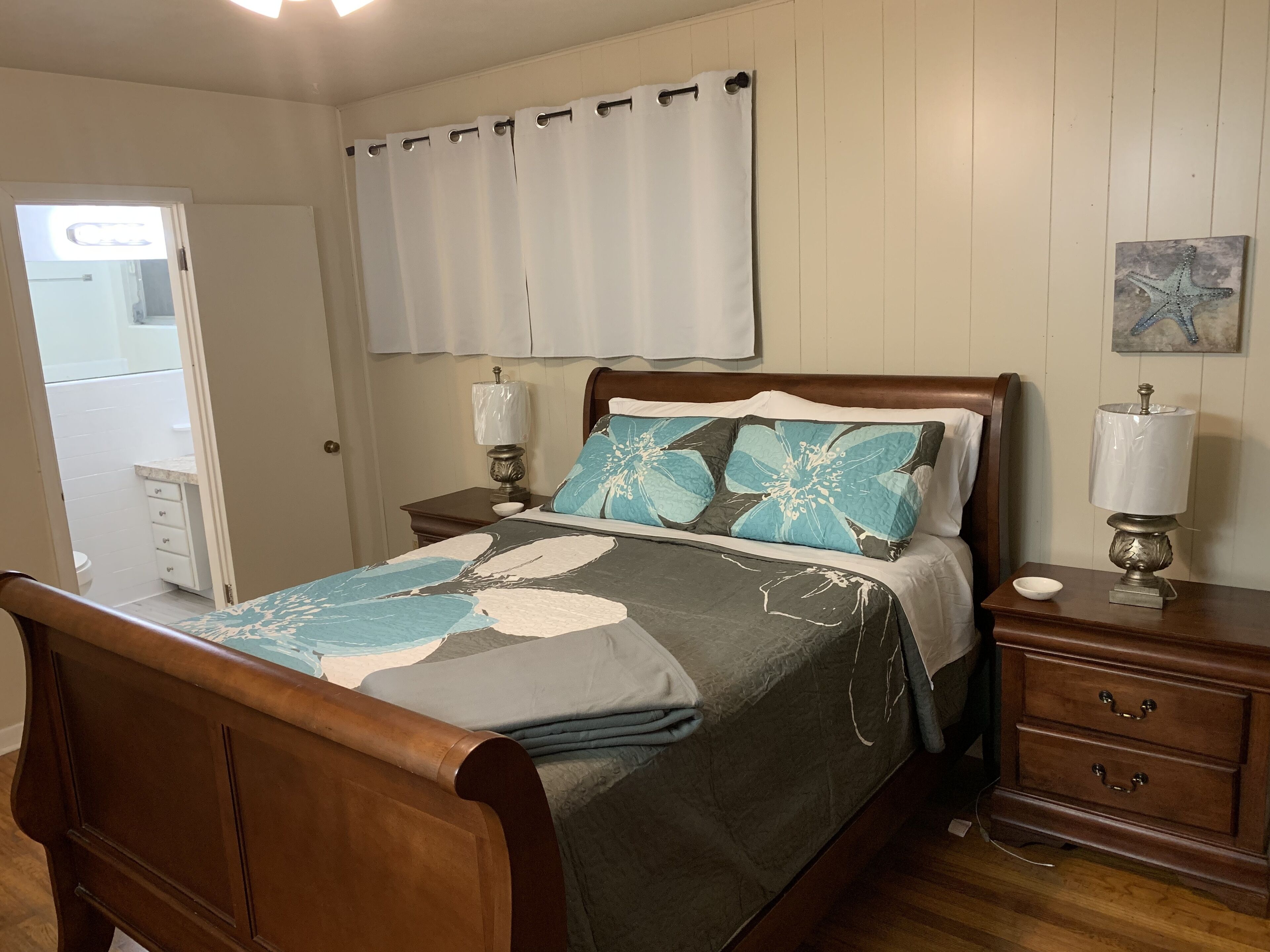 A bedroom with a wooden bed, a nightstand with a lamp, a dresser, and a bathroom visible through an open door.