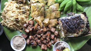 Variedad de platos sobre una hoja de plátano, incluyendo pescado a la parrilla, carne, verduras y un tazón pequeño de salsa.