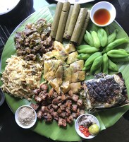 Variedad de platos sobre una hoja de plátano, incluyendo pescado a la parrilla, carne, verduras y un tazón pequeño de salsa.
