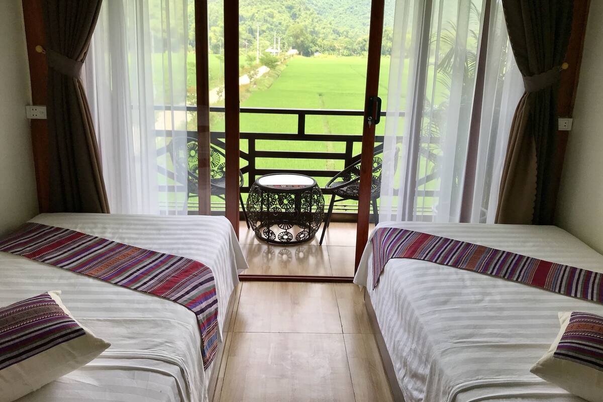 A room with two beds, a small table, and a view of a green landscape through sheer curtains.