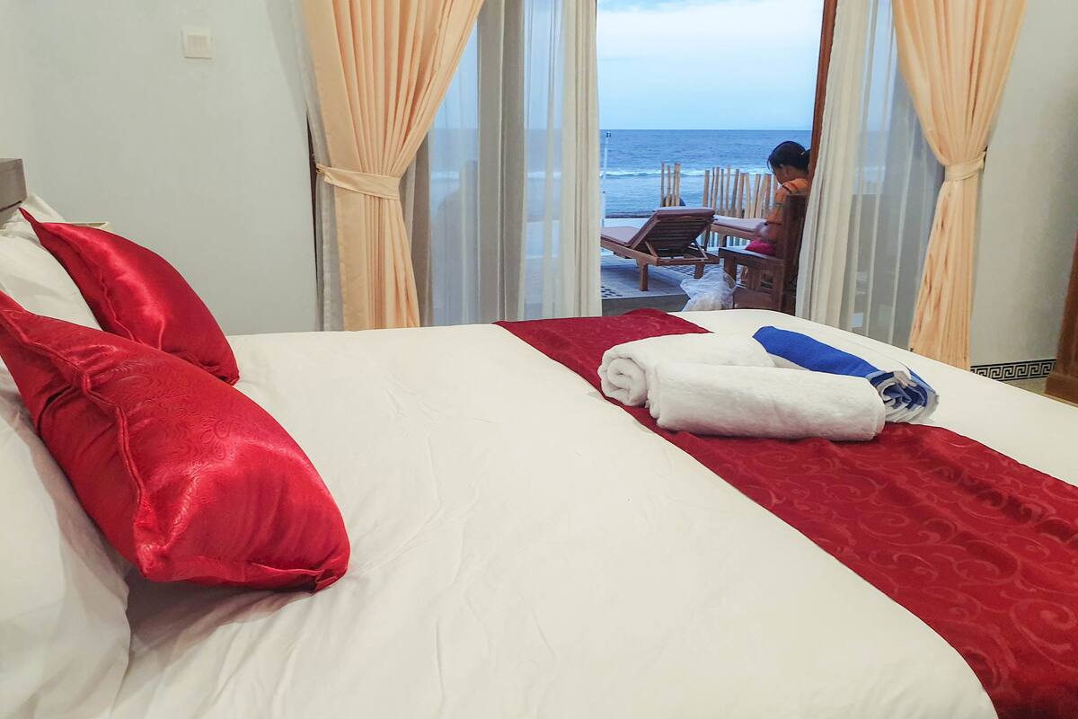 Quarto de hotel com uma cama, almofadas e manta vermelhas e brancas, e vista para o oceano através de grandes janelas.