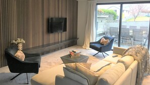 A modern living room with a flat-screen TV mounted on a wooden wall, a sofa, a coffee table with books, and a chair with a cushion.