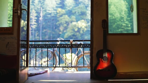 A room with a view of bicycles outside, a guitar leaning against the wall, and a door with a sign. - Nomads Hostel (Bhuntar)