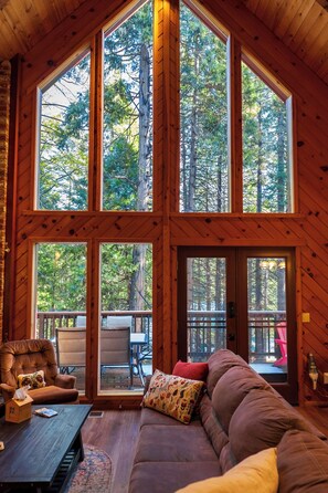 Una cabaña con ventanas amplias que ofrecen vistas a altos árboles. En el interior, hay un sofá de cuero marrón y una mesa de madera.