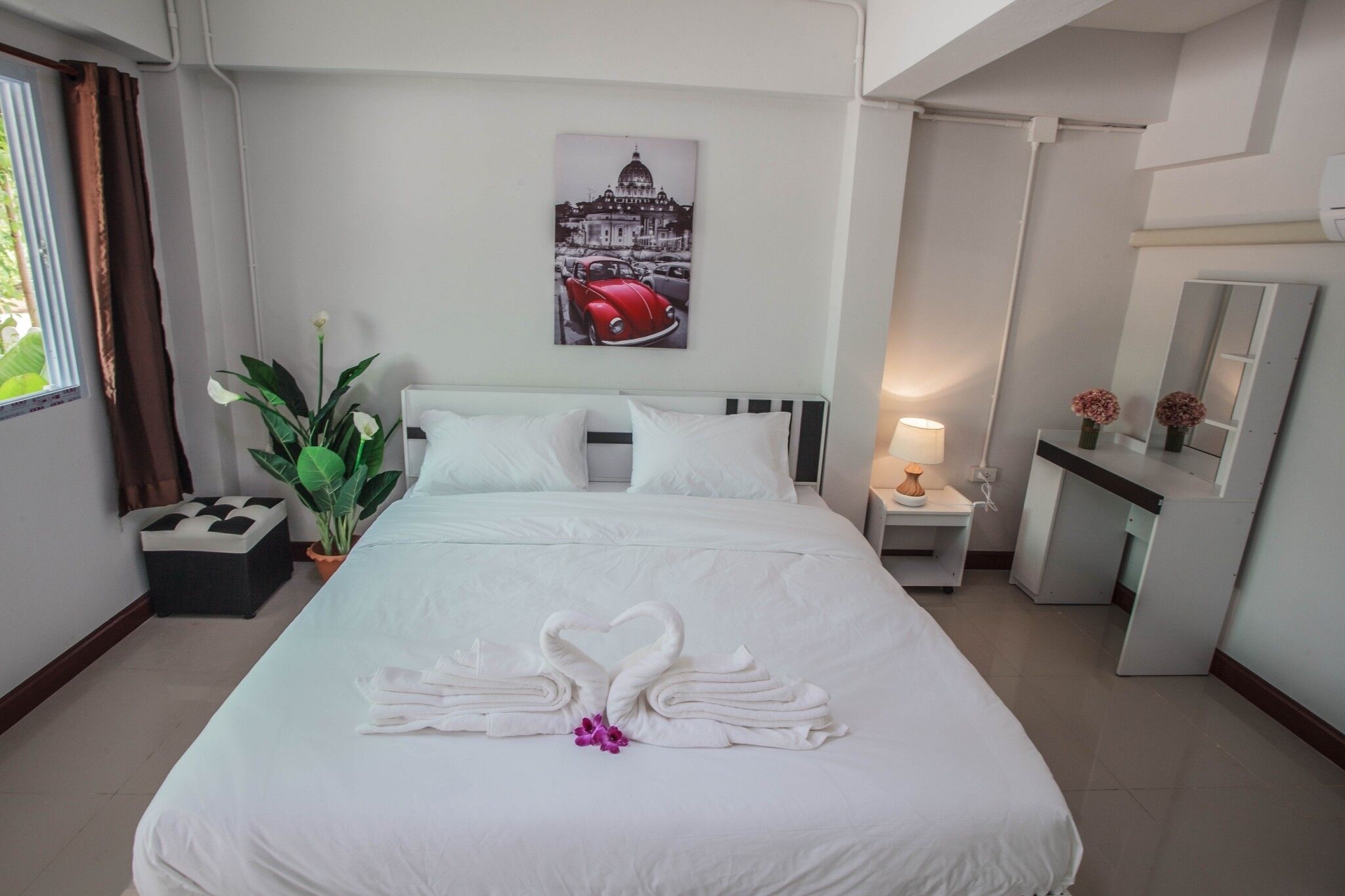 A neatly made bed with white linens and a heart-shaped towel arrangement, a potted plant, a nightstand with a lamp, and a wall-mounted picture of a car and building.