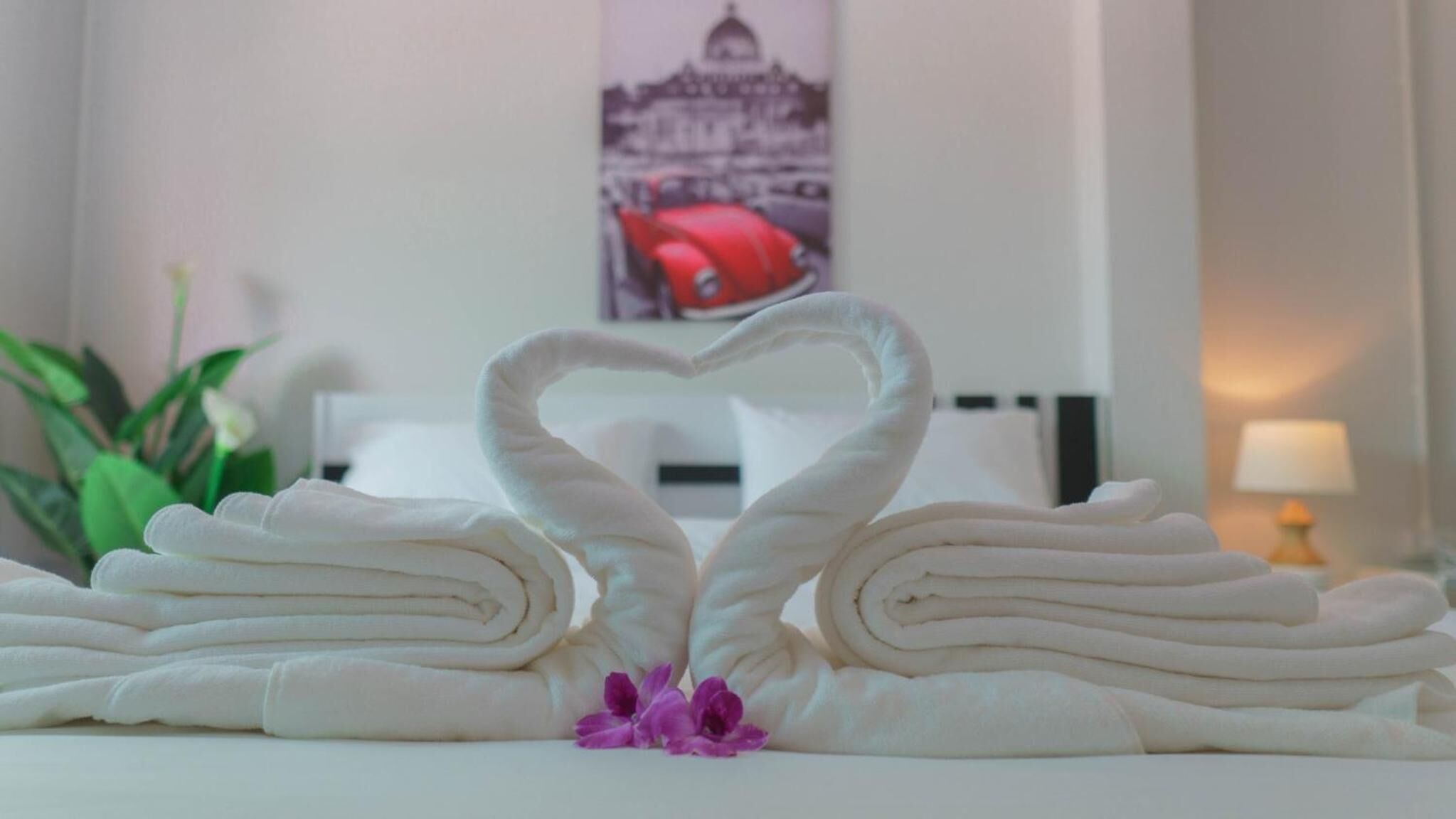 Una toalla blanca doblada con cuidado formando la forma de un corazón, con una flor morada en el centro, colocada sobre una cama en una habitación de hotel.