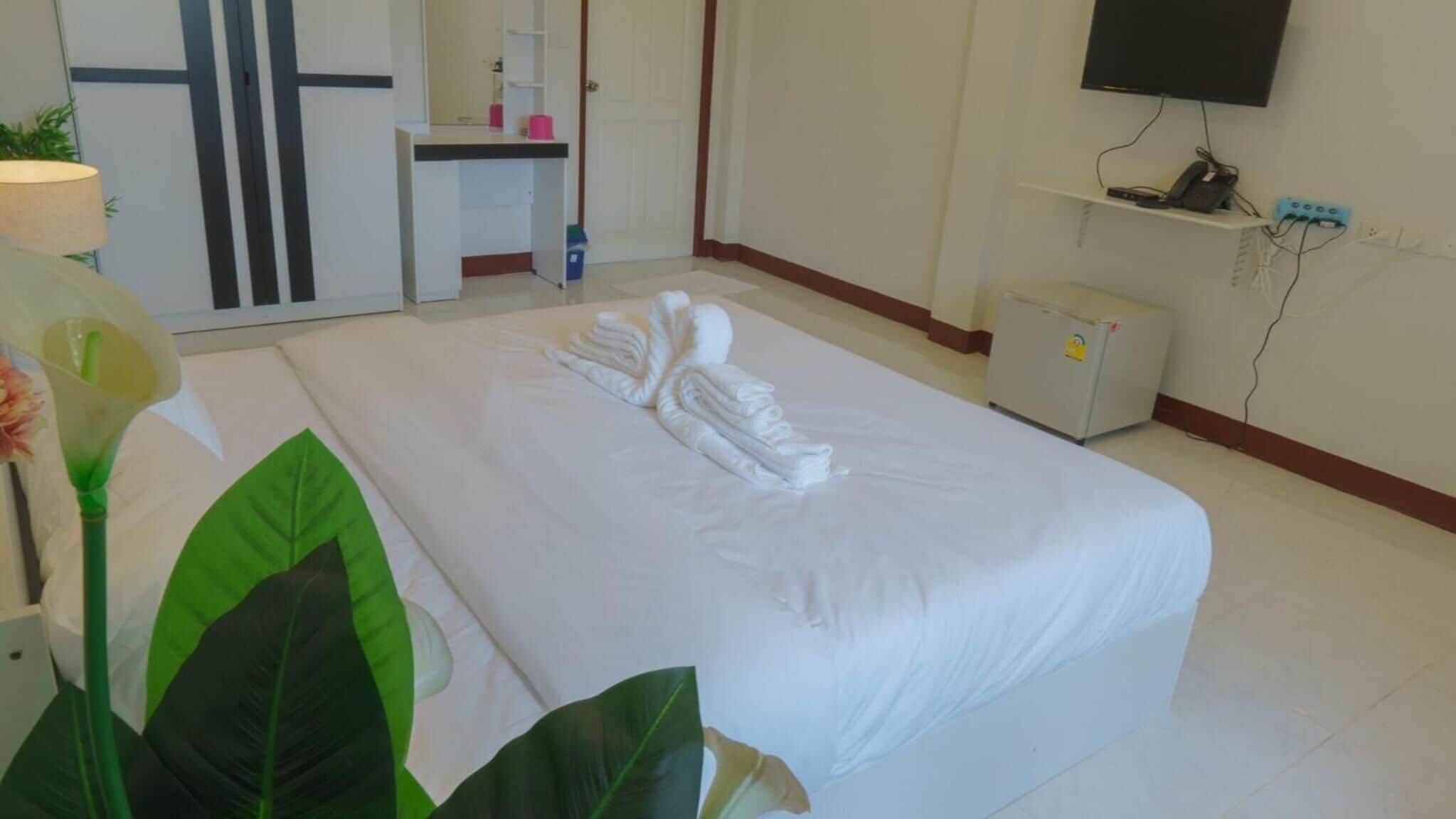Une chambre d’hôtel avec un lit bien fait, un bureau avec une chaise, un petit réfrigérateur, un téléviseur fixé au mur et une plante sur le côté gauche.