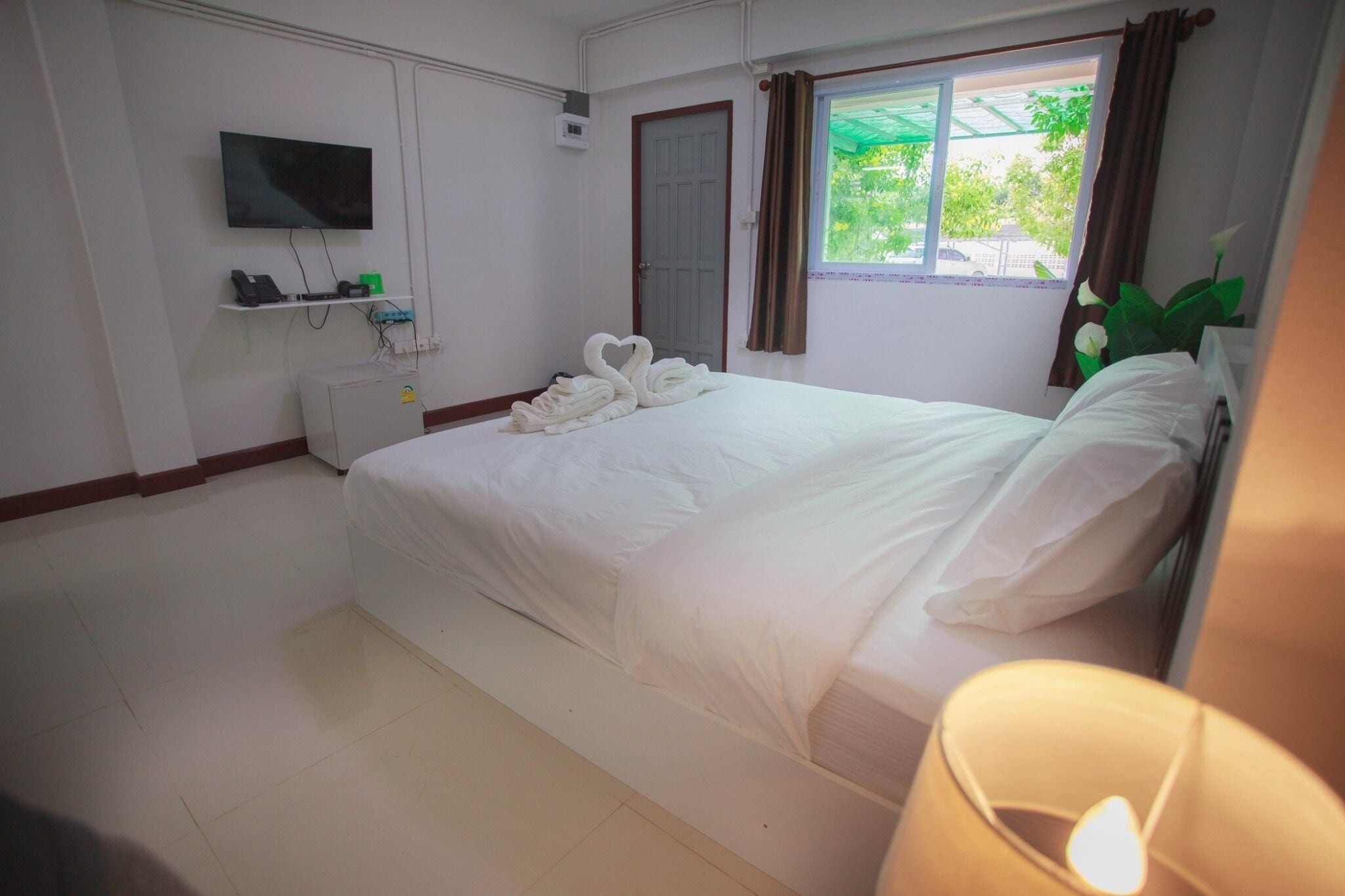 Habitación de hotel con cama, televisor montado en la pared, ventana con cortinas y una pequeña estantería con un teléfono y otros objetos.