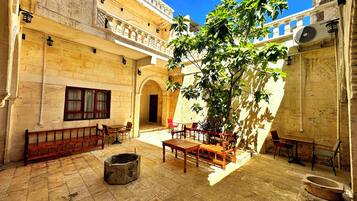 Un patio con suelo de piedra, bancos de madera, un árbol y un edificio tradicional con arcos y un balcón.