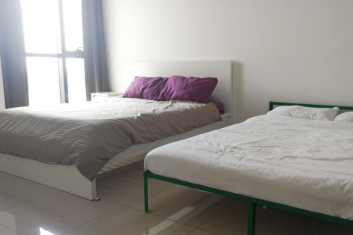 Two beds with white and purple bedding, a white nightstand, and a window with curtains.