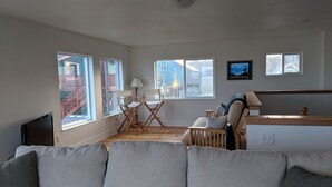 Un salon spacieux avec un grand canapé gris, une table basse en bois et un petit téléviseur posé sur un meuble. Plusieurs fenêtres laissent entrer la lumière naturelle et offrent une vue sur l’extérieur.