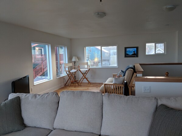 A spacious living room with a large gray sofa, a wooden coffee table, and a small TV on a stand. There are several windows providing natural light and views of the outside. - Ocean view house in Nye Beach (3B/2Ba) sleeps 8, by beach, shops & restaurants. (Newport)