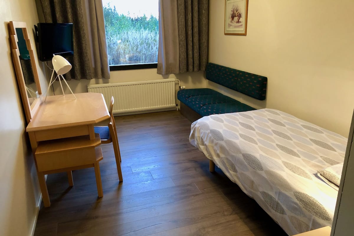 Quarto com escrivaninha de madeira, uma cadeira, cama com colcha estampada, janela com cortinas, radiador e quadro na parede.