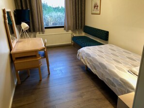 A bedroom with a wooden desk, a chair, a bed with a patterned bedspread, a window with curtains, a radiator, and a framed picture on the wall.