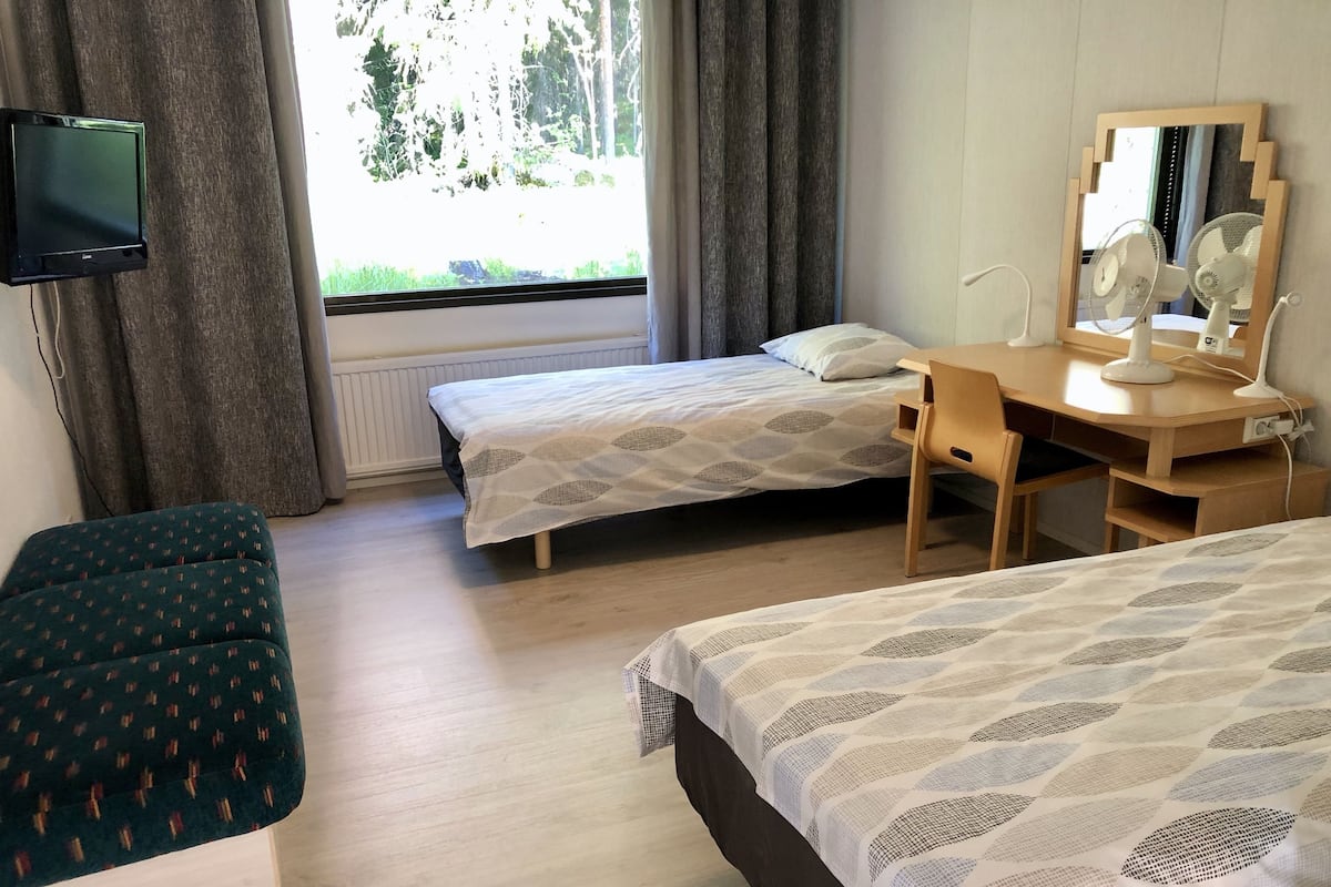 Quarto com duas camas de solteiro, uma escrivaninha com espelho, uma cadeira, televisão e janela com cortinas.