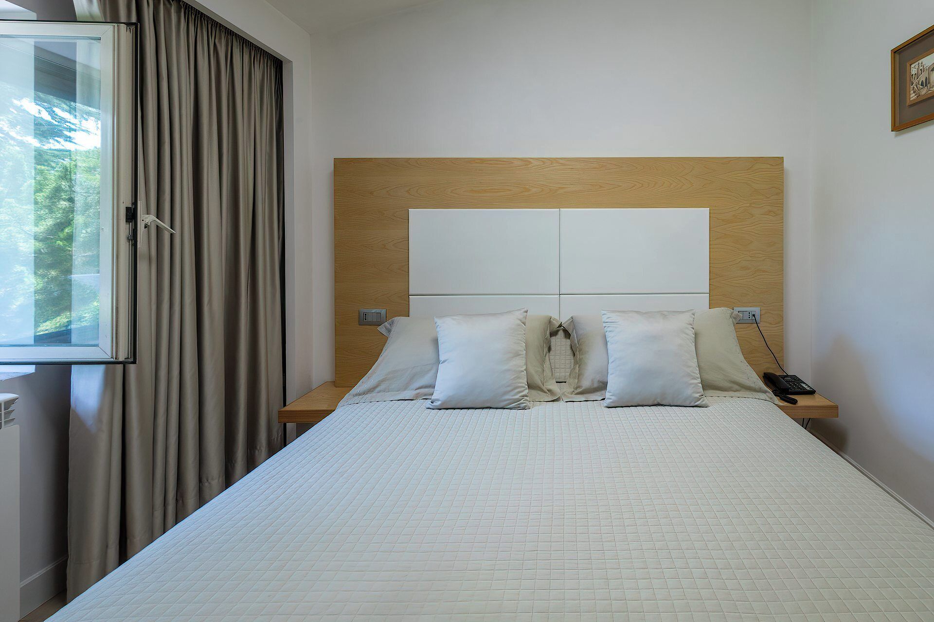 Habitación de hotel con una cama grande, dos mesitas de noche, una ventana con cortinas y un cabecero de madera.