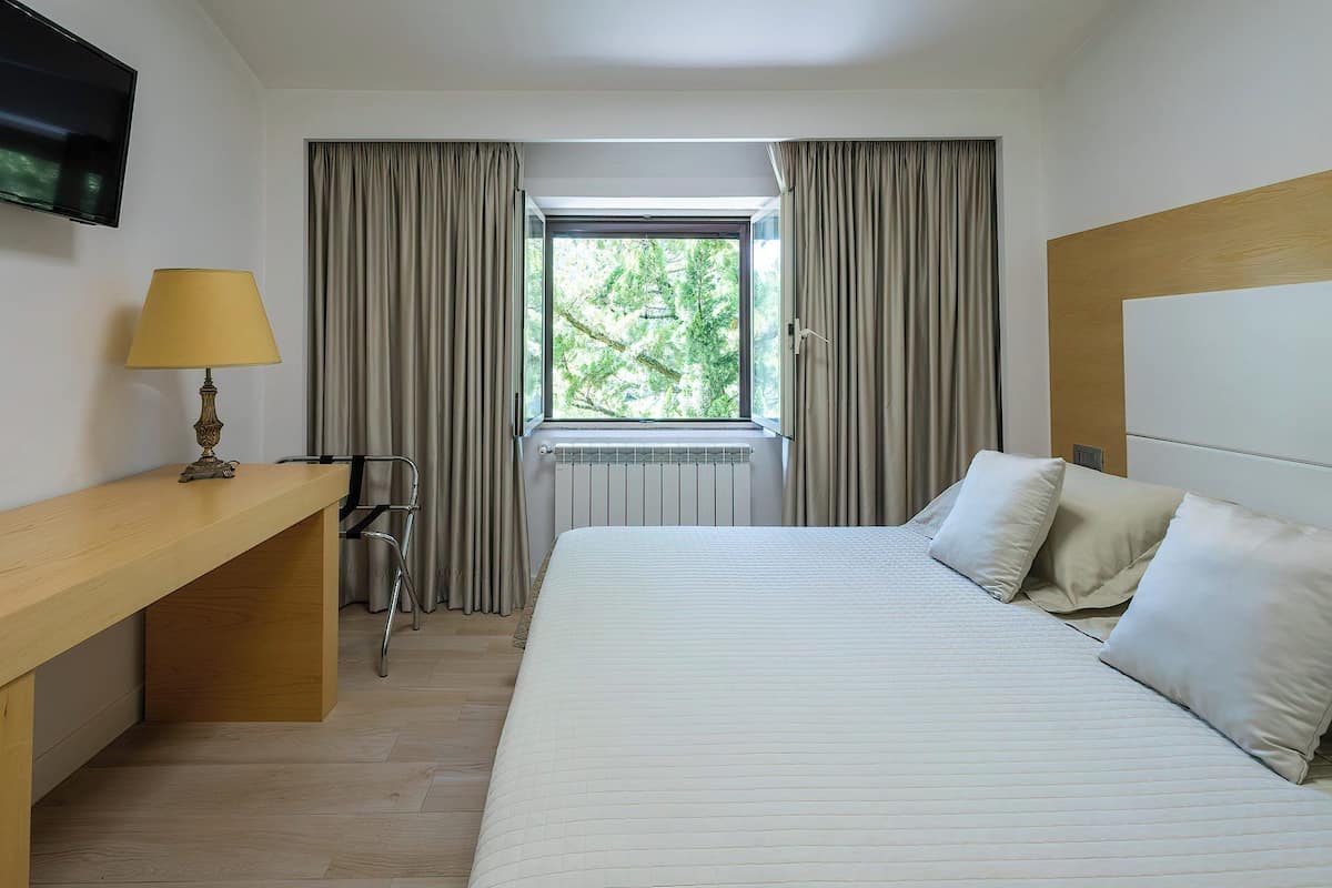 Une chambre d’hôtel avec un lit, un bureau, une chaise, une lampe et une fenêtre avec des rideaux.