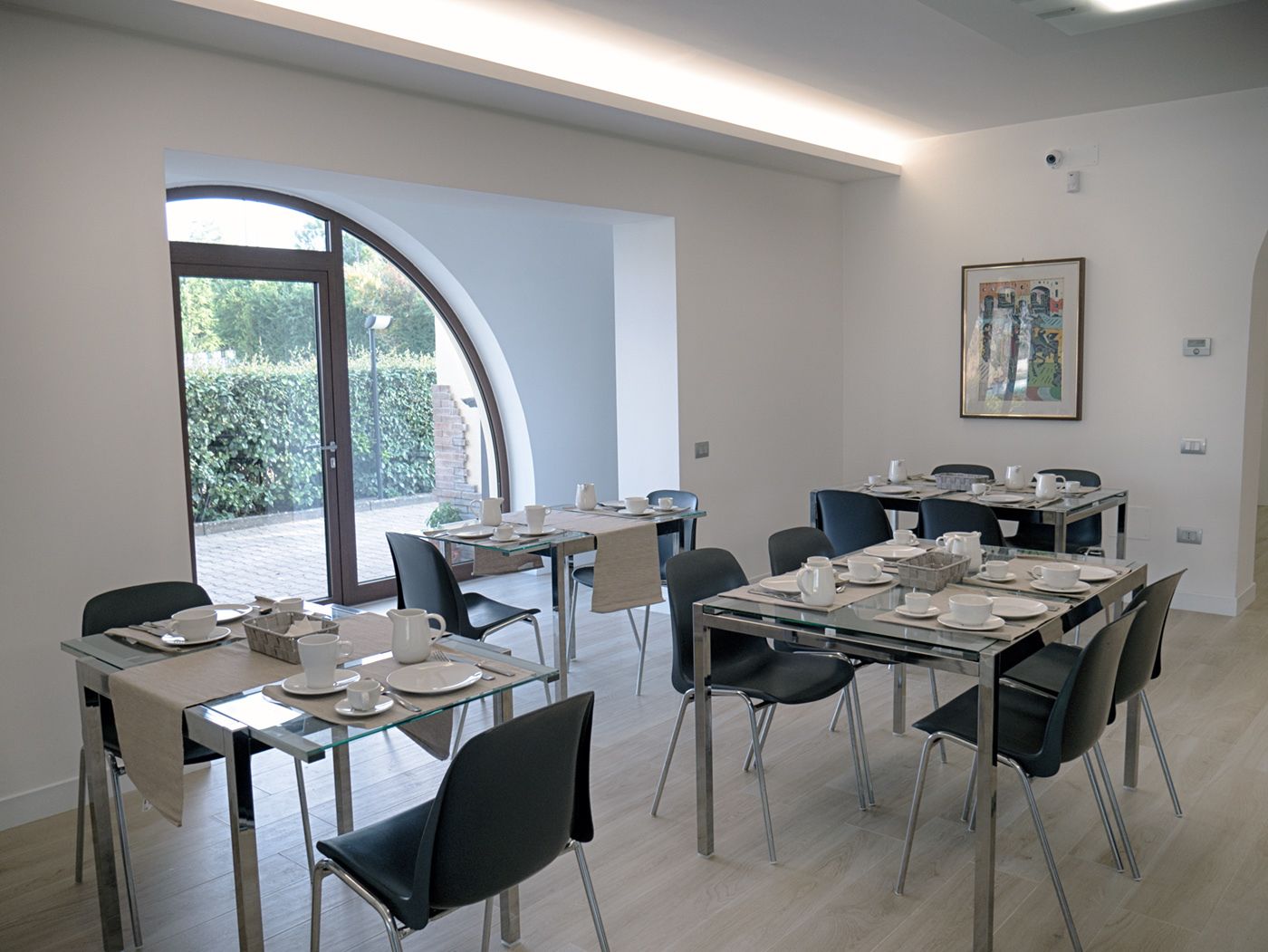 Un comedor moderno con mesas y sillas de vidrio, un ventanal con vistas a la vegetación y una obra de arte enmarcada en la pared.