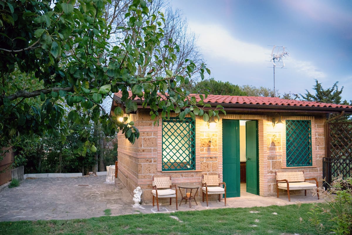 Un petit bâtiment en pierre avec un toit en tuiles, deux fenêtres encadrées de vert et une porte. On y trouve deux chaises et une petite table à l’extérieur.