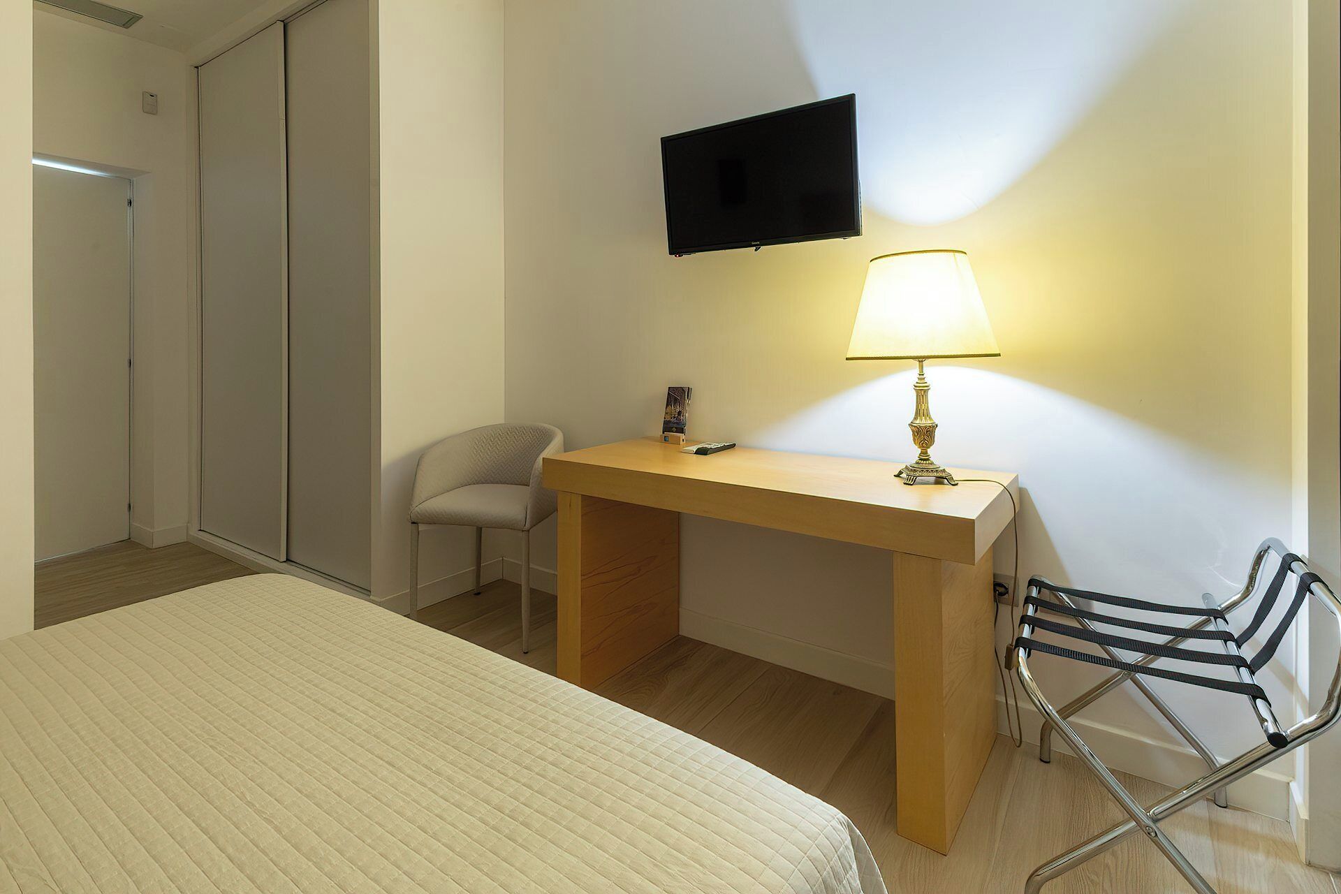 A hotel room with a bed, a desk with a lamp, a chair, a TV mounted on the wall, and a wardrobe.